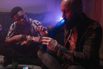 Two men sitting, playing music, using bong