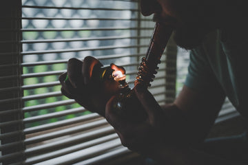 Man lighting and smoking a glass bong beside a window